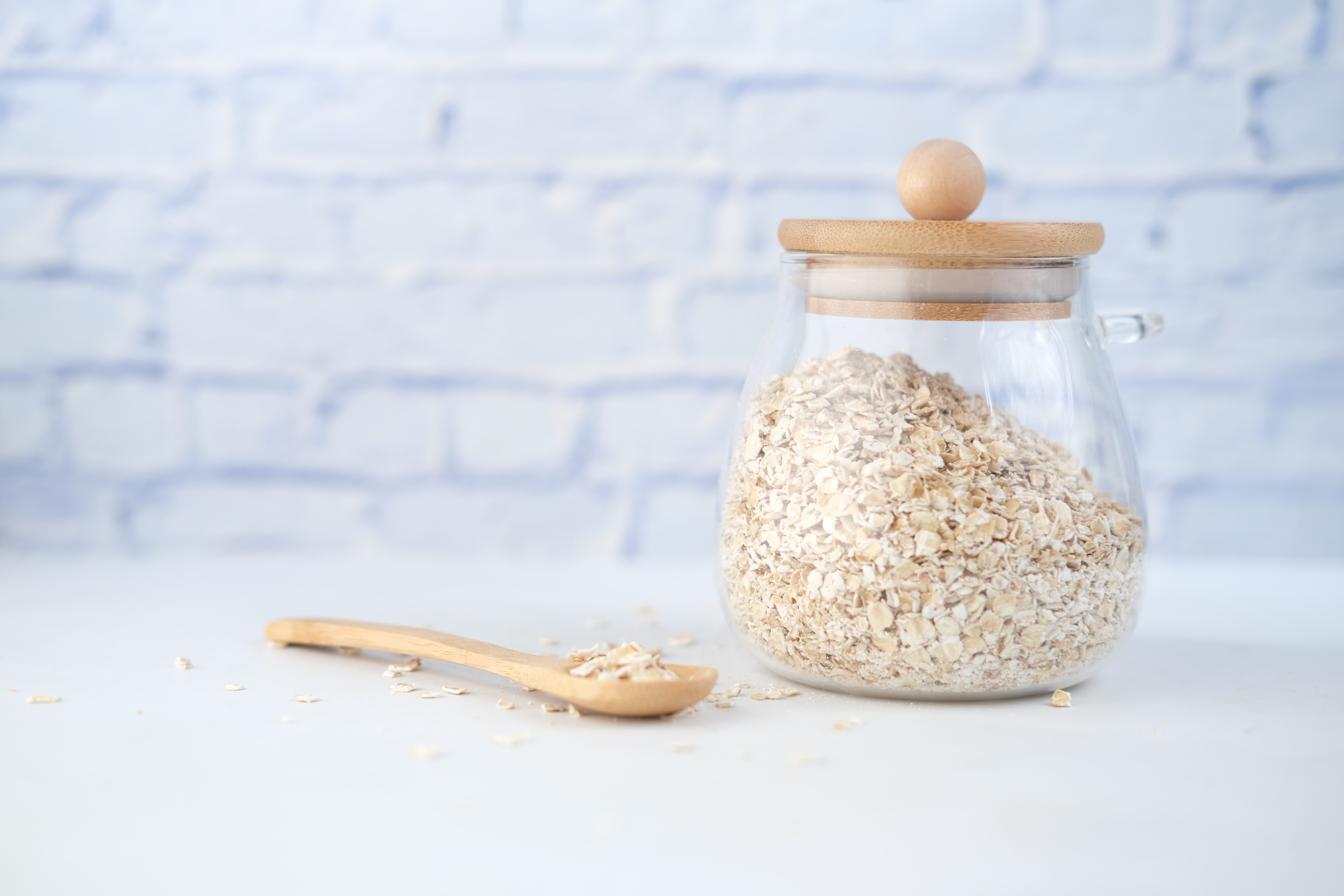 Beta-Glucan Wirkung: Hier erfährst du alles Wissenswerte zum Super-Ballaststoff weißer Tisch vor weißer Ziegelwand, darauf ein ovales Glas mit Holzdeckel gefüllt mit Gerstenflocken, daneben Gerstenflocken auf einem Holzlöffel