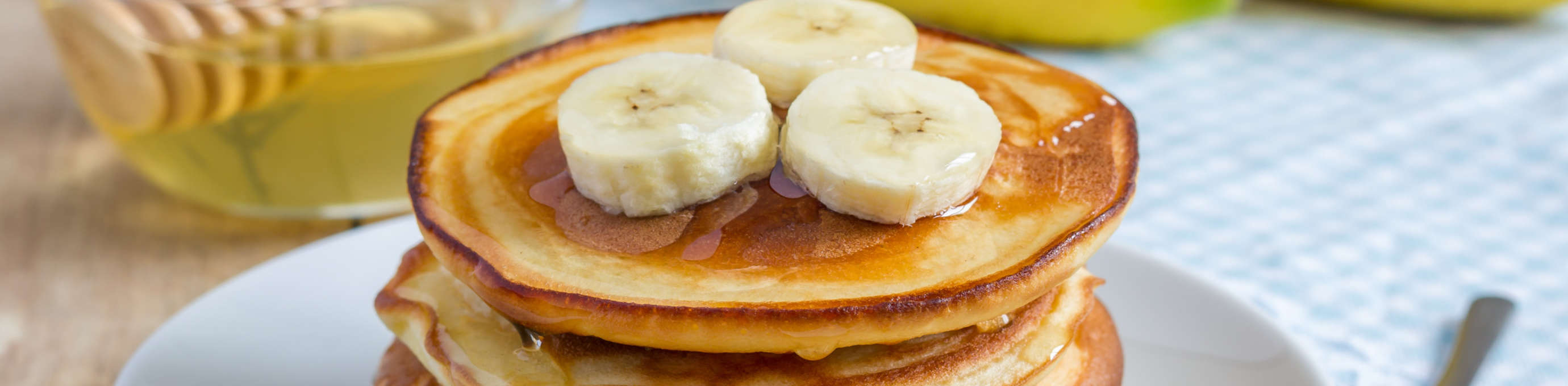 Gestapelte Pancakes aus Gerstoni Gerstenflocken mit Honig überzogen und mit Bananenscheiben garniert, liegend auf weißem Teller