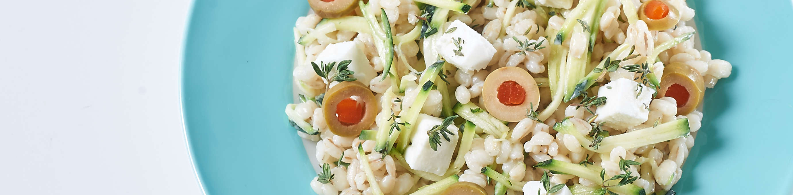 Salat aus Gerstoni Gourmet Gerste mit Zucchini, Feta und Olivenscheibchen auf hellblauem Teller