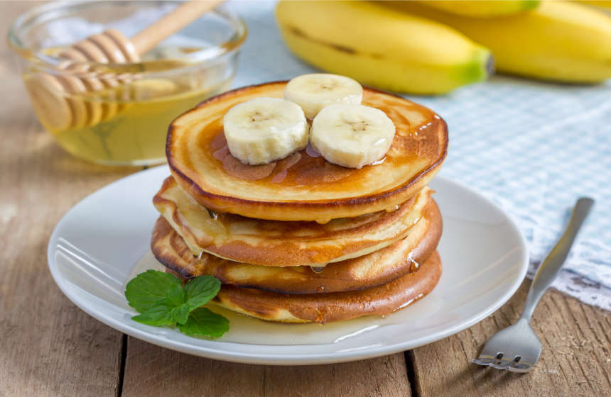 Gestapelte Pancakes aus Gerstoni Gerstenflocken mit Honig überzogen und mit Bananenscheiben garniert, liegend auf weißem Teller